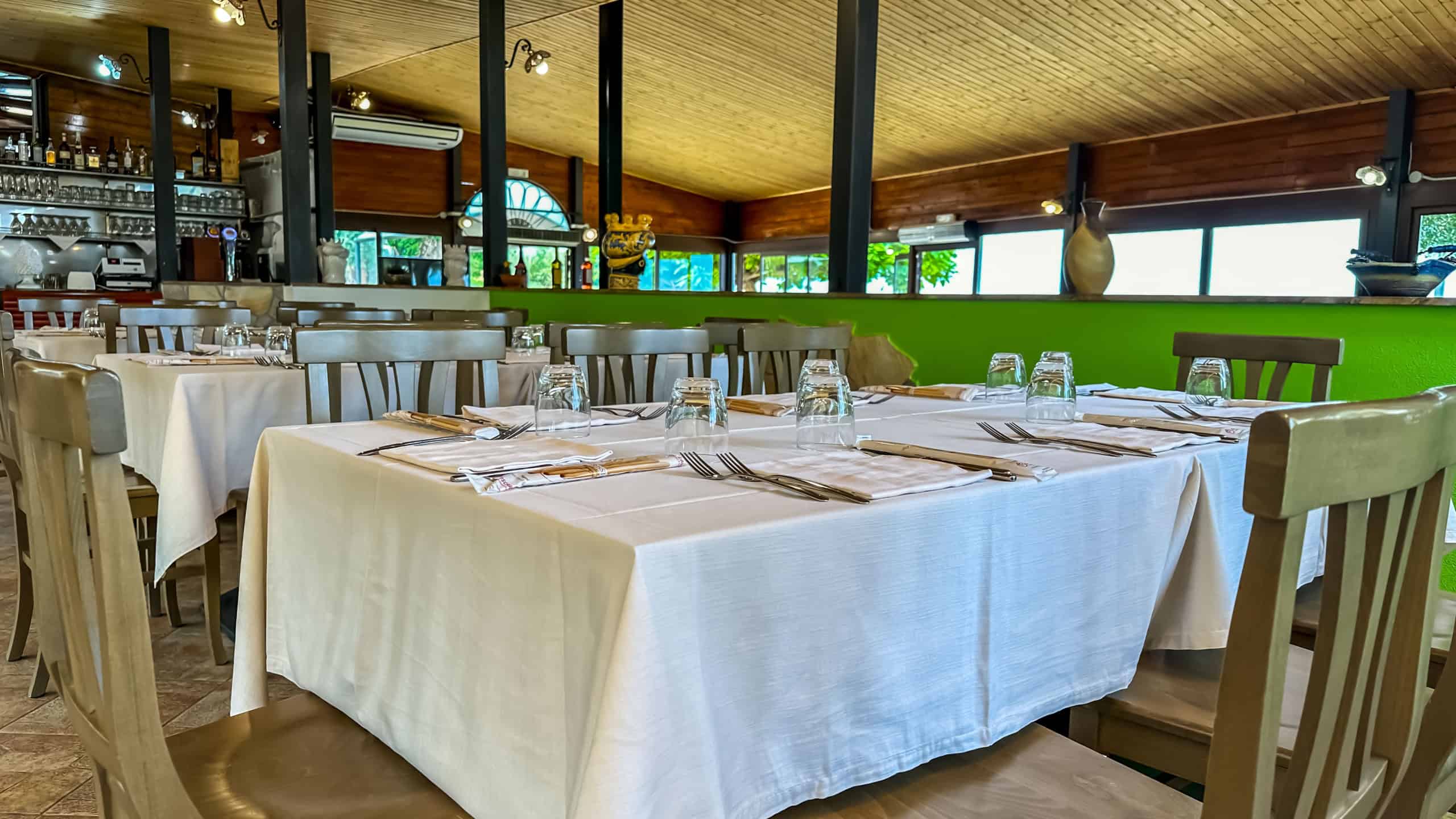 Tavolo elegante in ristorante con tovaglia bianca, posate e bicchieri pronti per i clienti, ambiente accogliente con arredi in legno e grandi finestre che lasciano entrare molta luce naturale.