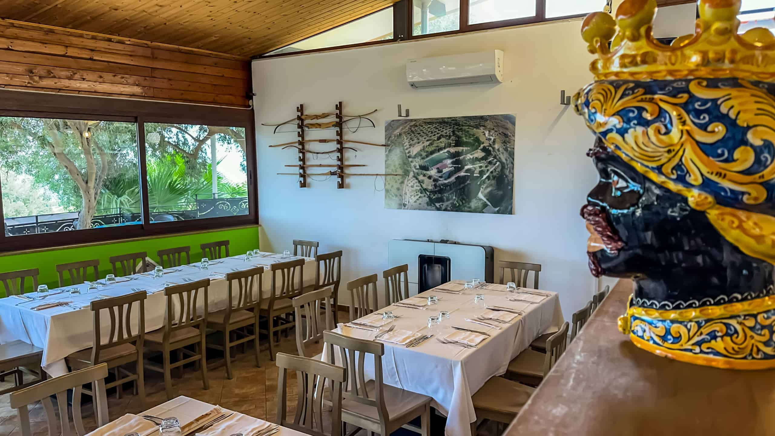 Sala da pranzo accogliente con grandi finestre che offrono vista sul verde, tavoli eleganti con tovaglie bianche e sedie in legno, decorazioni artistiche e un grande drago decorativo in primo piano.