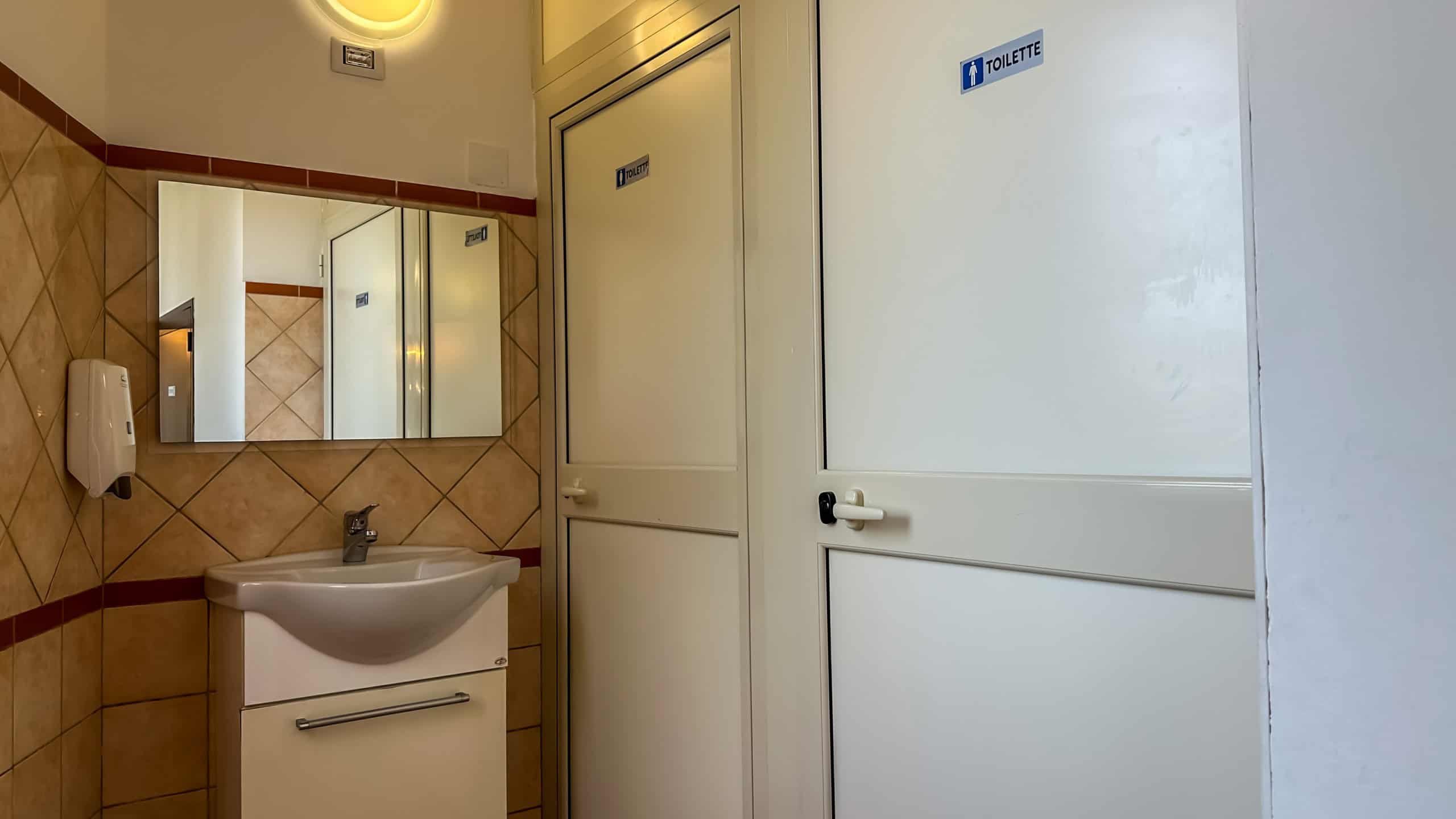 Lavandino e porta del bagno pubblico con segnale "Toilette" in un ambiente interno.