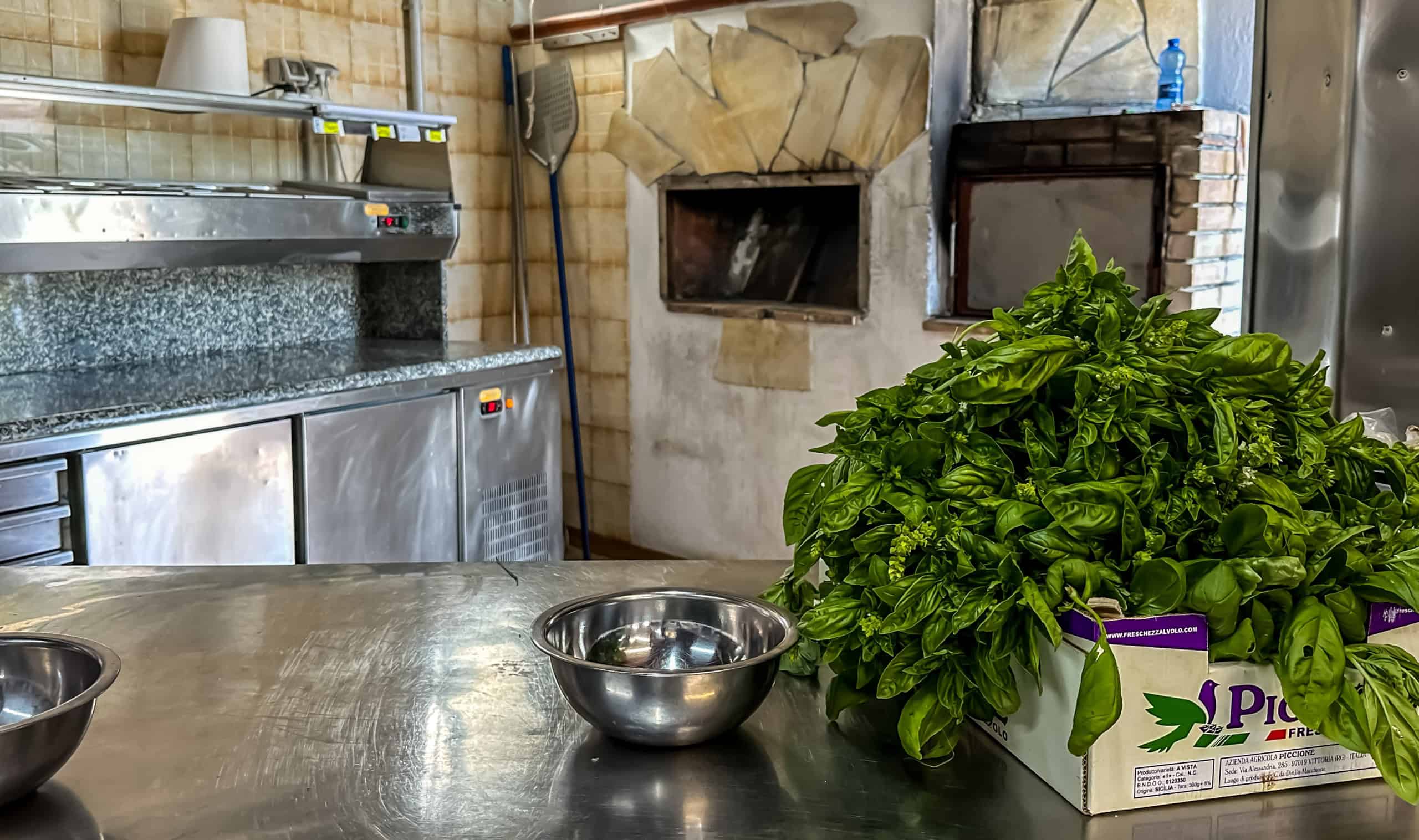 Pane fresco e basilico in un ambiente di cucina con forno a legna e attrezzature in acciaio inox, ideale per preparare piatti tradizionali italiani e pizza fatta in casa. Atmosfera rustica e accoglien.