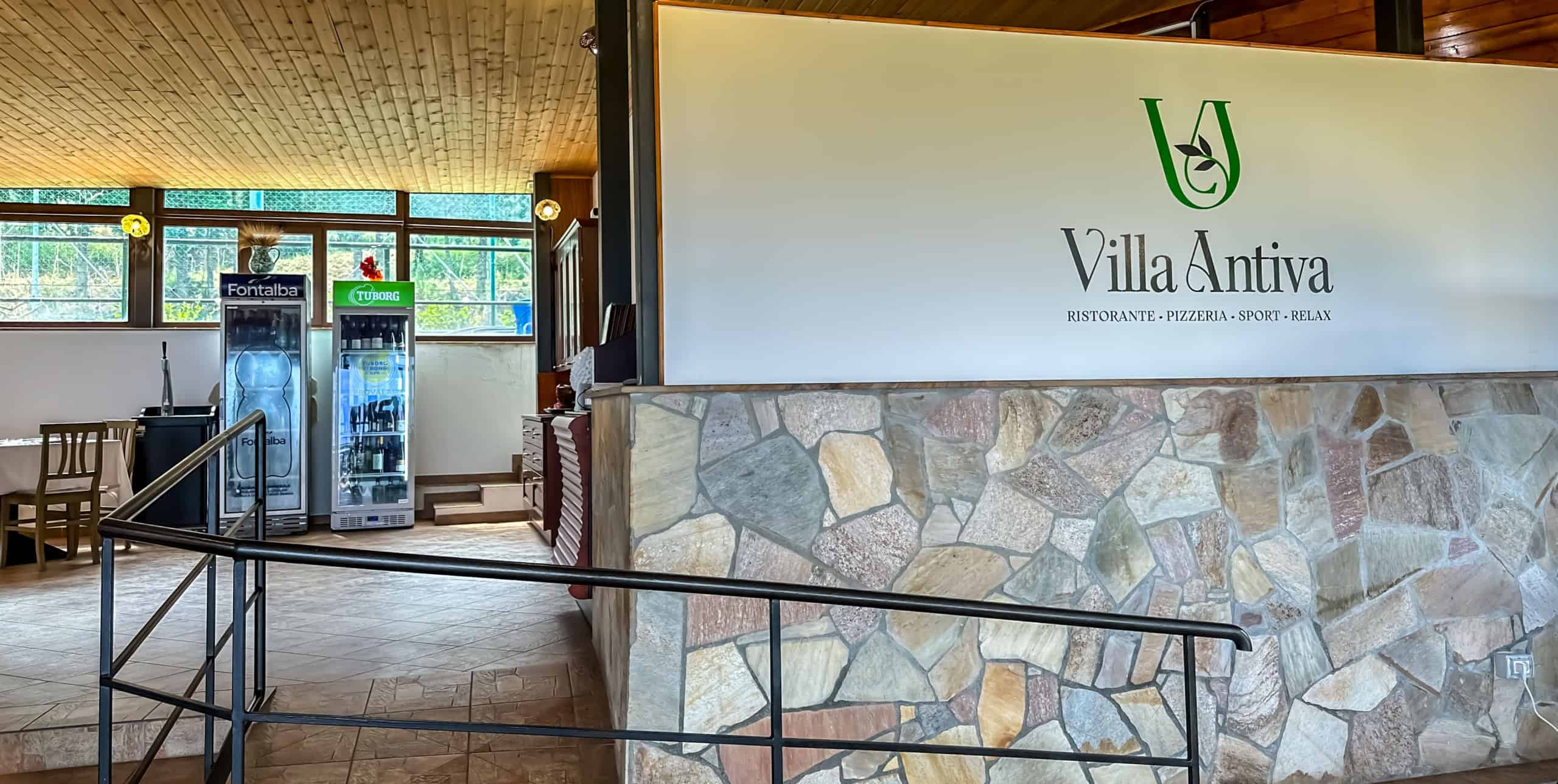Pannello con il logo Villa Antiva, ristorante e pizzeria, all’interno di un locale accogliente con pareti in pietra e grandi finestre che lasciano entrare molta luce naturale.