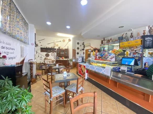 Interno di un bar o caffetteria con tavoli, sedie e un bancone con prodotti alimentari e bevande, atmosfera accogliente e luminosa.