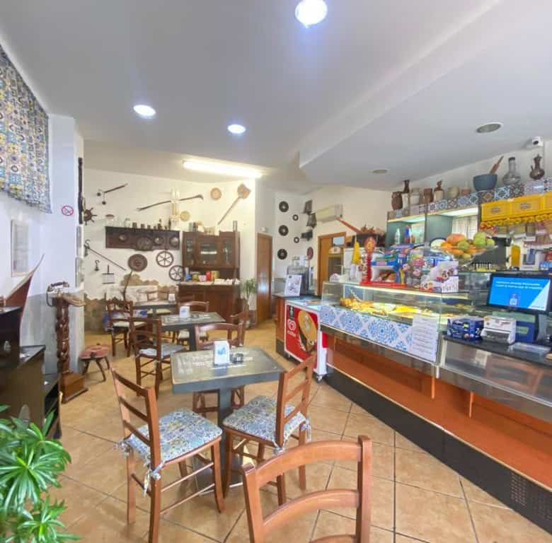 Interno di un bar o caffetteria con tavoli, sedie e un bancone con prodotti alimentari e bevande, atmosfera accogliente e luminosa.