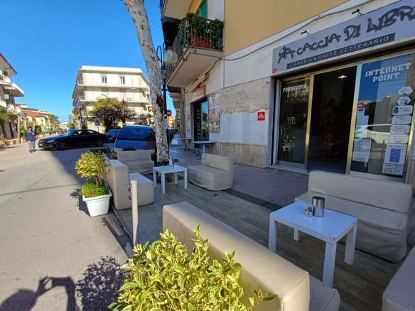 Un angolo di strada con sedute all'aperto e un negozio di libreria con caffetteria, in una zona urbana soleggiata e vivace, ideale per momenti di relax e socializzazione all'aperto.