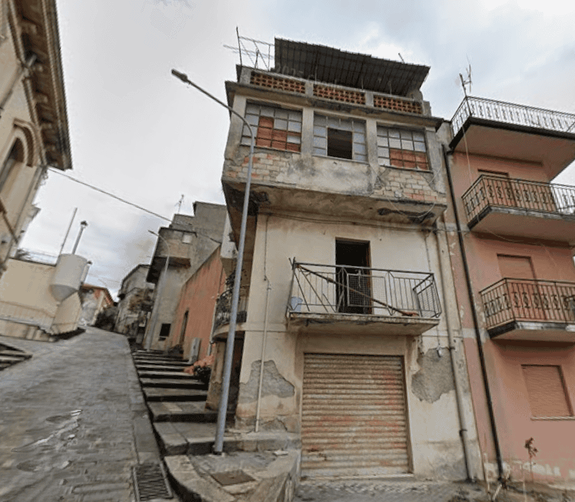 Edificio residenziale su strada in pendenza con balcone e tetto solare, in zona urbana con altri edifici vicini, in condizioni di deterioramento.