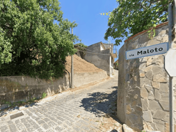 Ampia strada in salita con ciottoli e muri in pietra, segnaletica stradale via Maloto, circondata da alberi verdi e cielo azzurro, in un quartiere tranquillo e pittoresco.