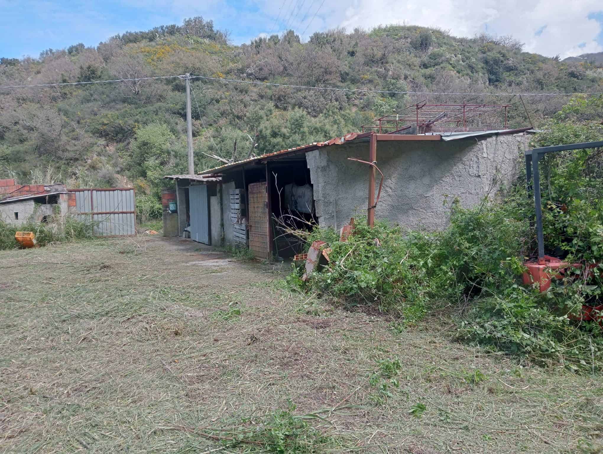 Vecchio capanno in legno e cemento in campagna, con vegetazione circostante e sfondo collinare, in condizioni di abbandono e deterioramento.