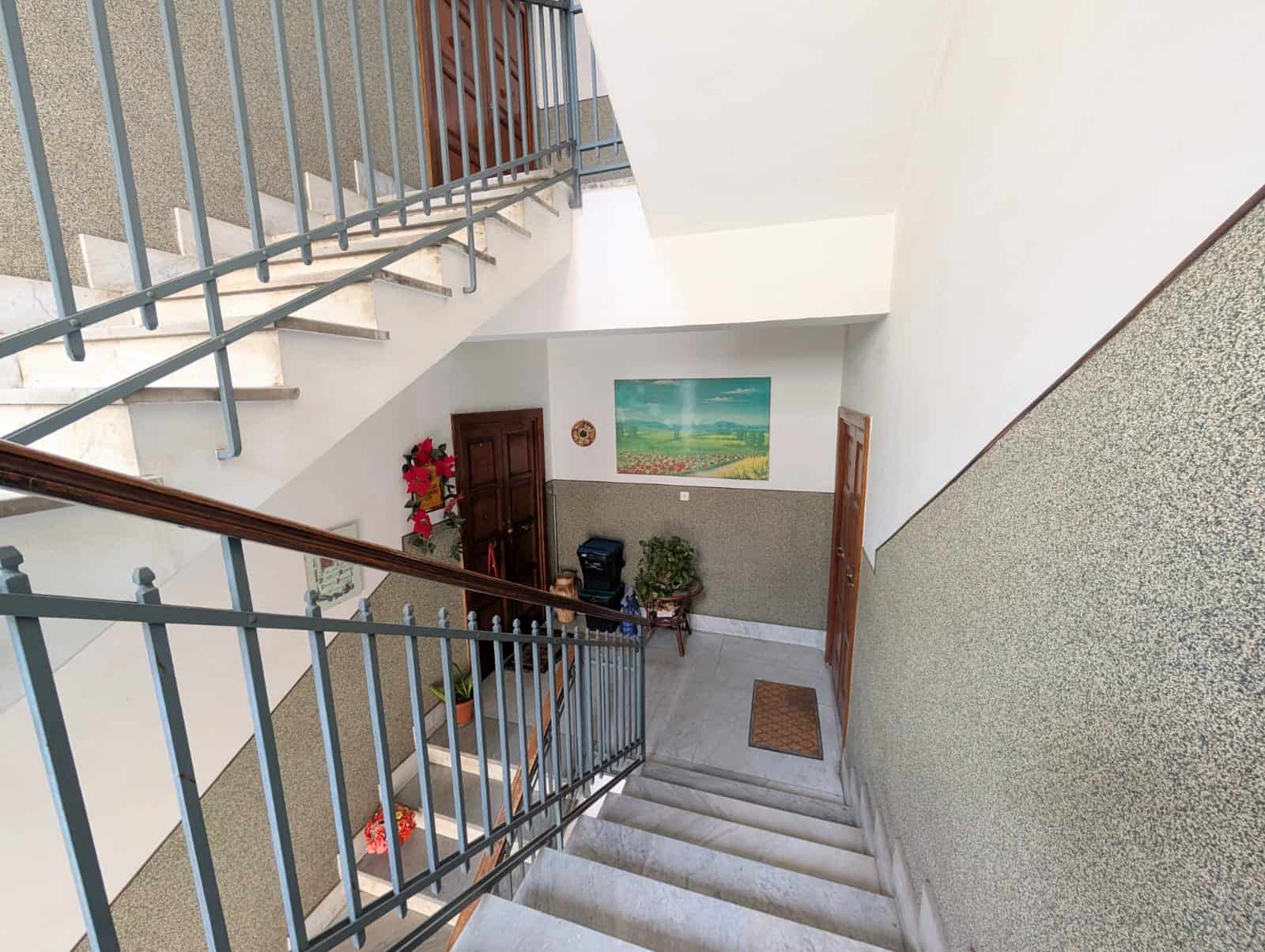 Escalier interno di un edificio residenziale con ringhiera in metallo, pareti bianche e decorazioni floreali, zona di ingresso con tappetino e quadro paesaggistico alle pareti.