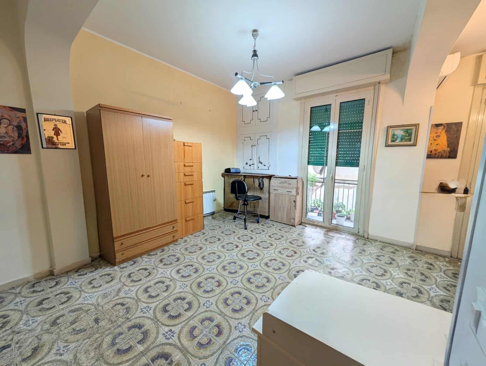 Ampio soggiorno con scrivania, armadio e accesso a balcone, ideale per vivere e lavorare in casa, con pavimento decorato e illuminazione centrale.