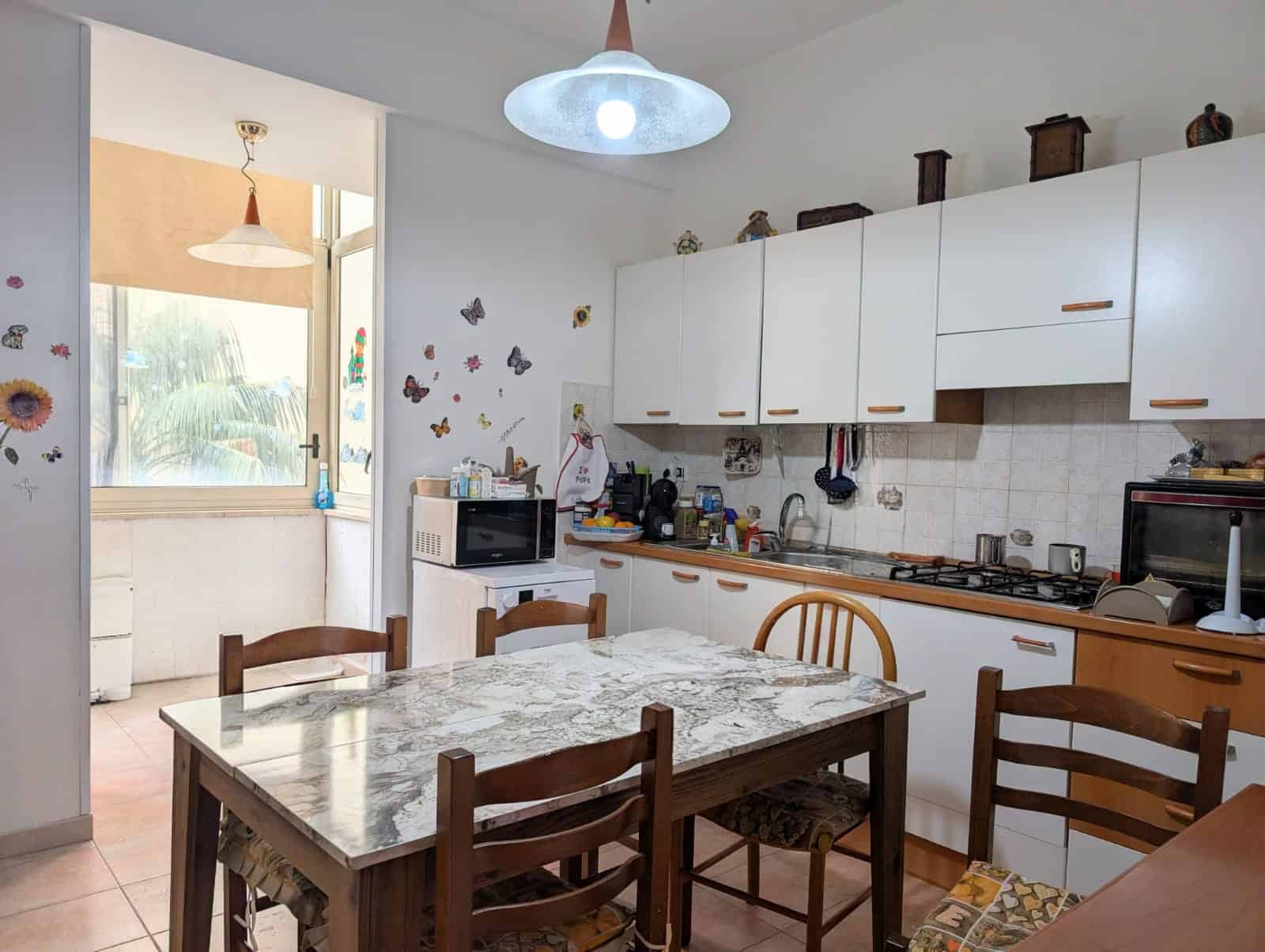 Pannello di cucina moderna con mobili bianchi, tavolo in marmo e sedie in legno, ambiente luminoso e accogliente, ideale per famiglie e appartamenti in affitto o vendita.