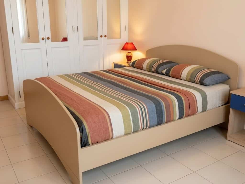 Letto matrimoniale con testiera imbottita in una camera da letto luminosa, decorata con lampada da tavolo e armadio a muro bianco, ambiente accogliente e moderno.
