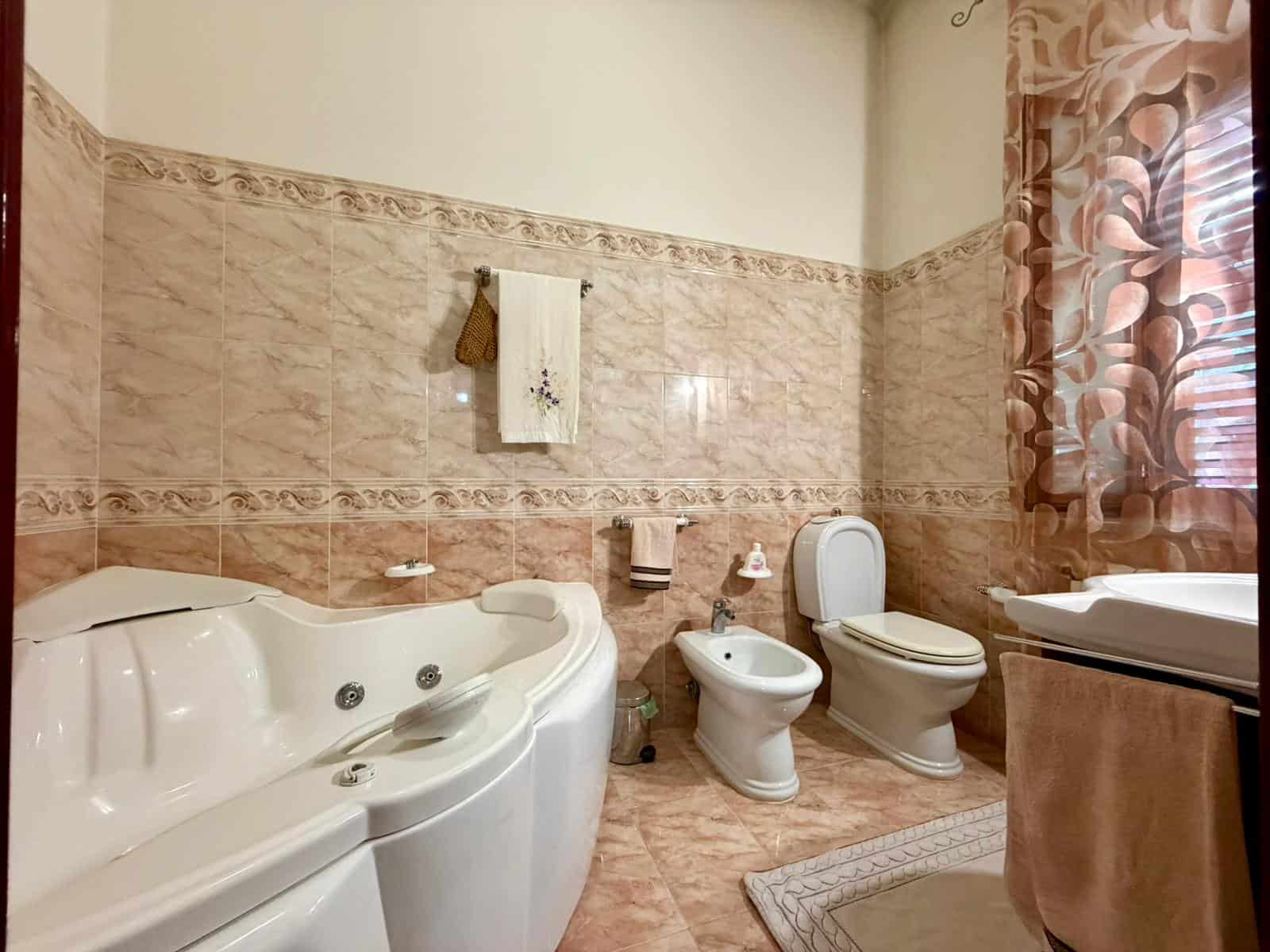 Acqua calda e idromassaggio in bagno spazioso con vasca idromassaggio, WC e bidet, illuminato da luce naturale attraverso una finestra con tende floreali, ideale per relax e benessere.