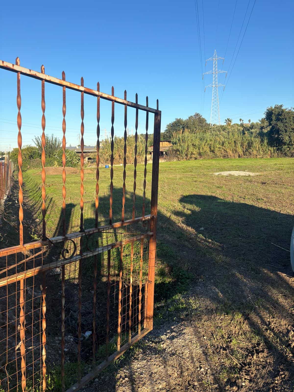 Cancello in ferro battuto rustico aperto su un'area verde all'aperto con cielo sereno, ideale per proprietà in vendita o in affitto, con elementi naturali e infrastrutture di energia elettrica visibil.