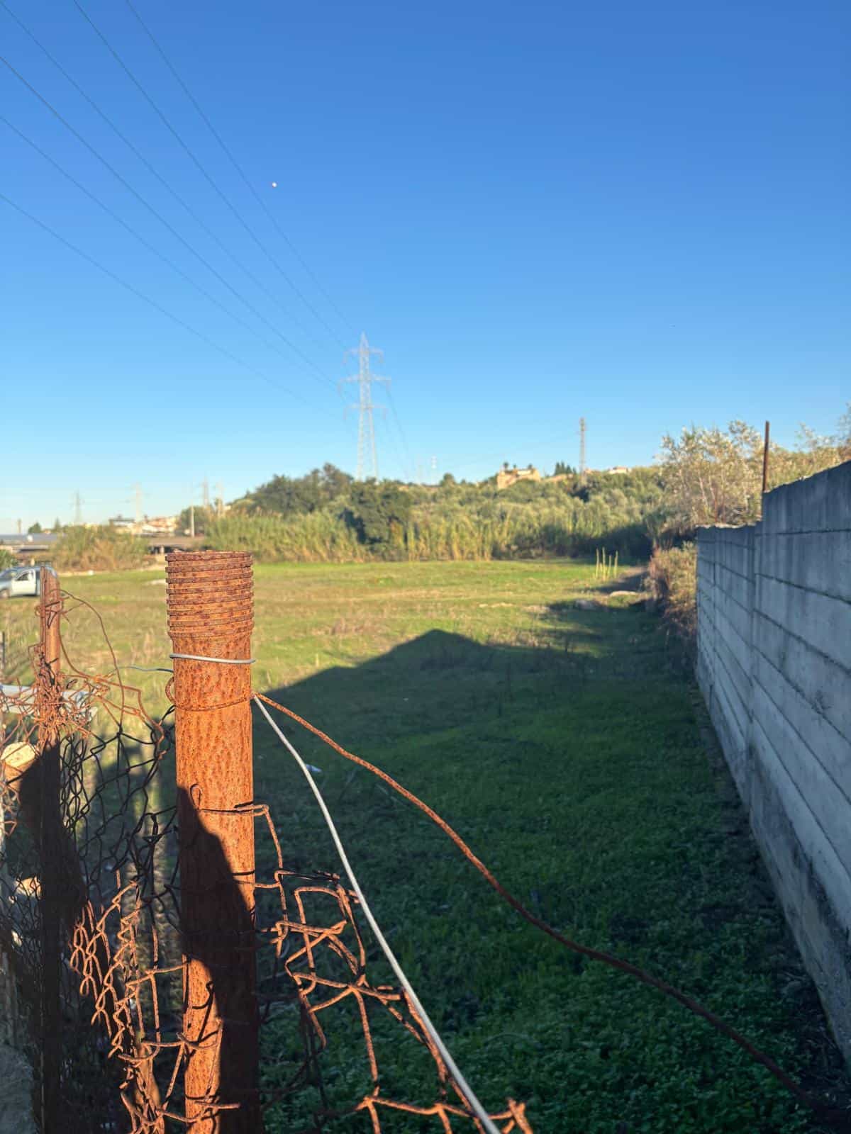 Ampia area di terreno verde con recinzione in ferro arrugginita e cielo azzurro, ideale per progetti di sviluppo immobiliare o giardinaggio, vicino a infrastrutture e natura.