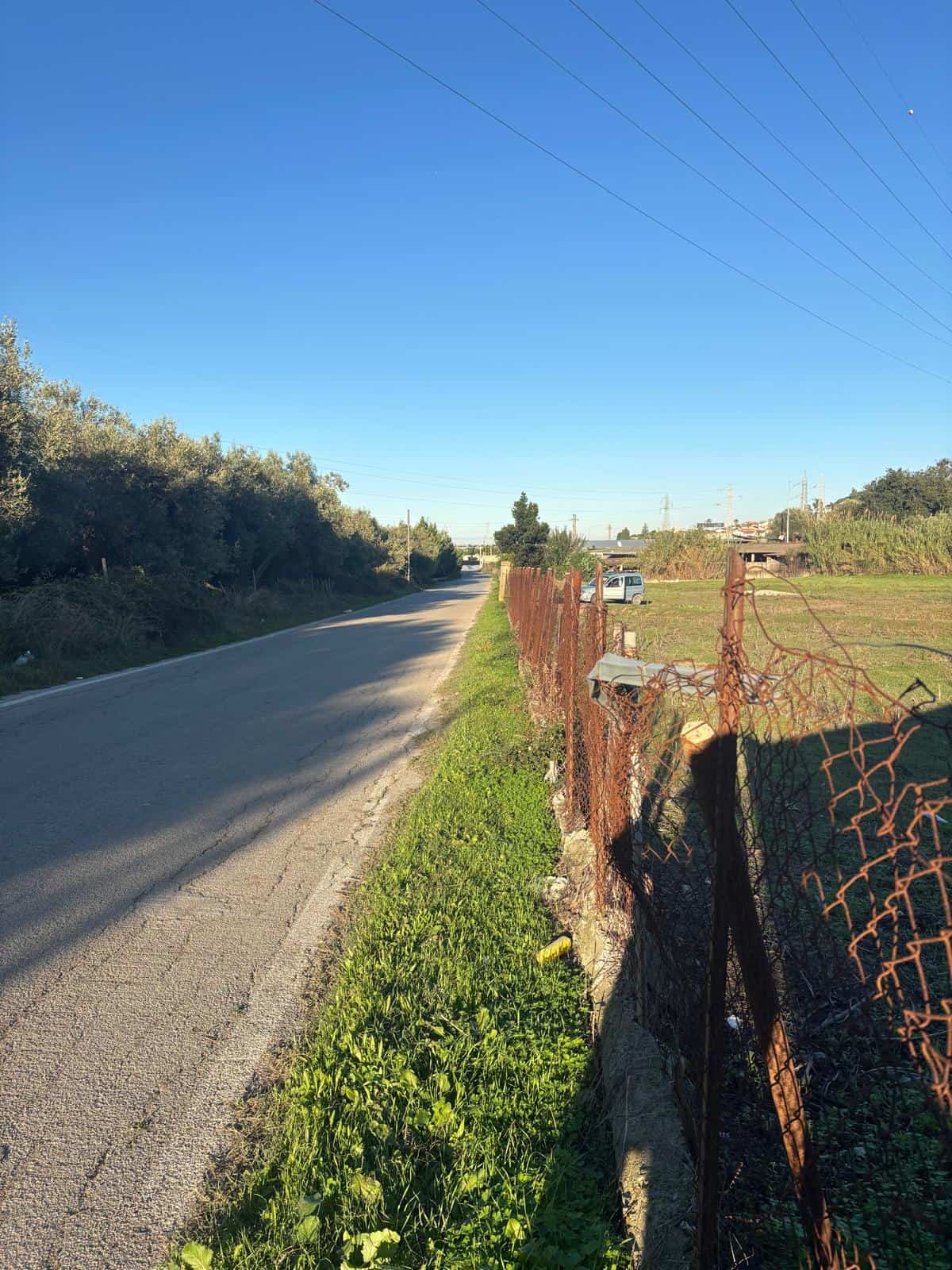 Finestra di strada rurale con recinzione in ferro arrugginito, cielo sereno e vegetazione verde lungo il ciglio, in un paesaggio tranquillo e naturale.