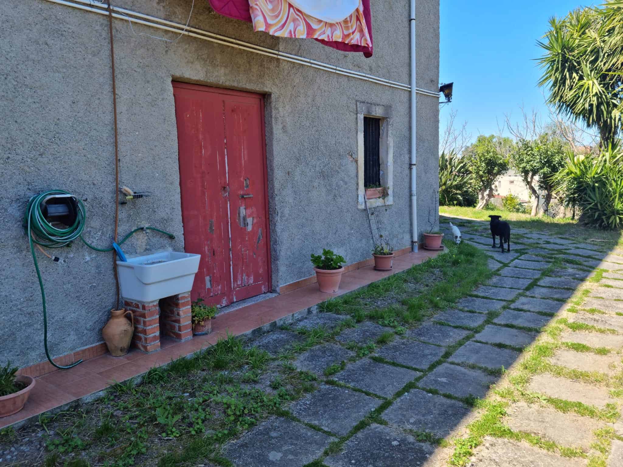 Porte esterne in legno rosso con parete in cemento, giardino con piante e sentiero in pietra, ambiente esterno di una casa rurale italiana.