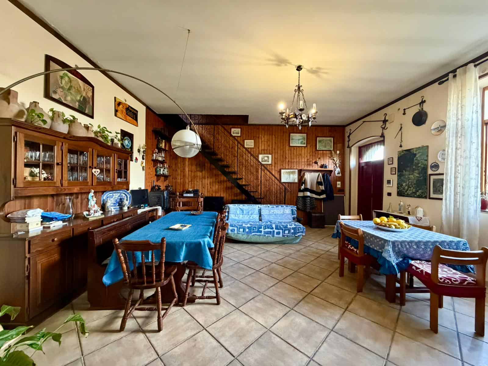 Interno di una casa con arredi tradizionali, tavoli con tovaglie blu, divano e decorazioni vintage, ambiente accogliente e luminoso.