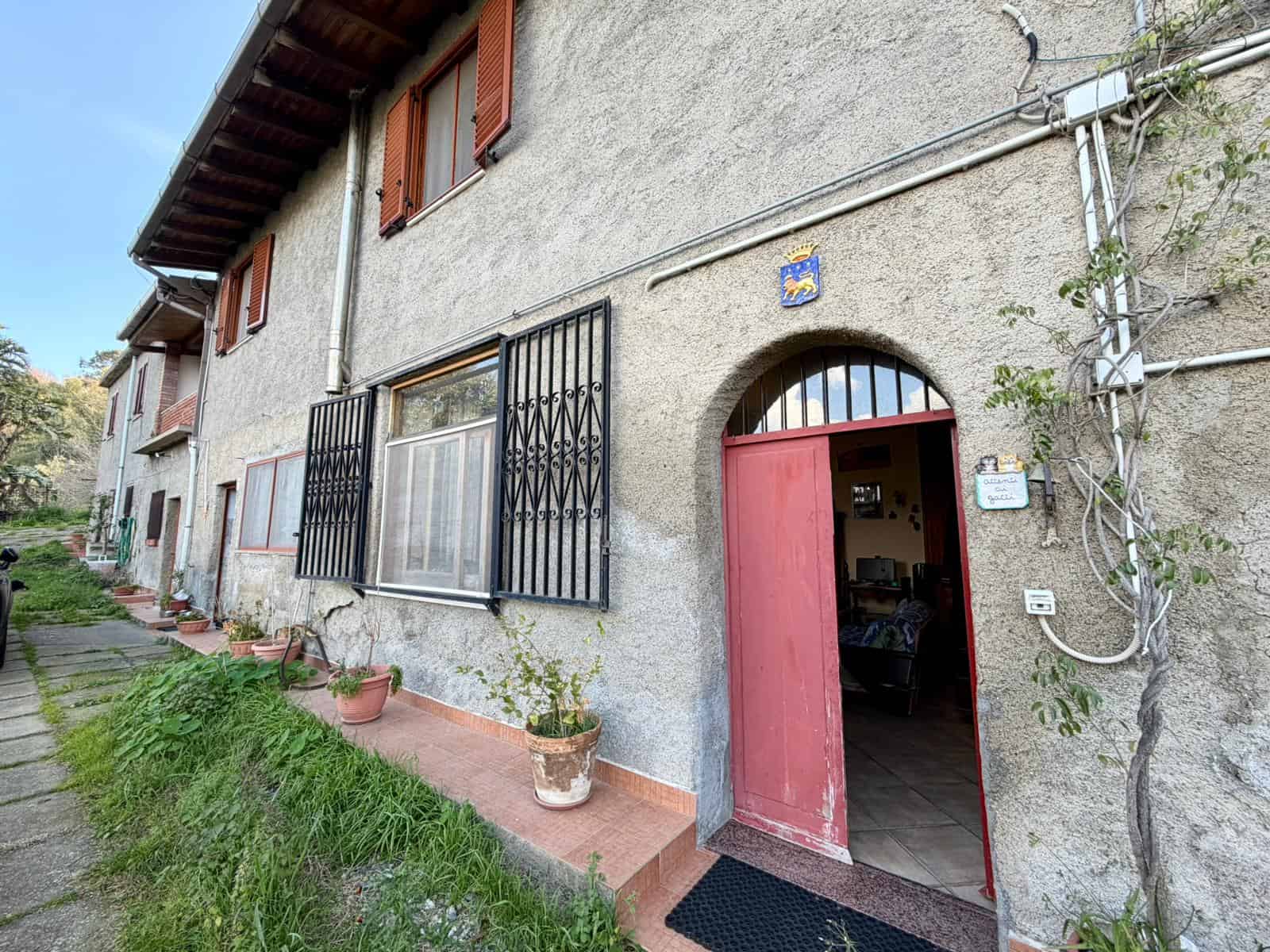 Cancello d'ingresso di una casa rustica con pareti in pietra, finestre con grate di sicurezza e piante in vaso, in un contesto di zona residenziale tranquilla e verde.