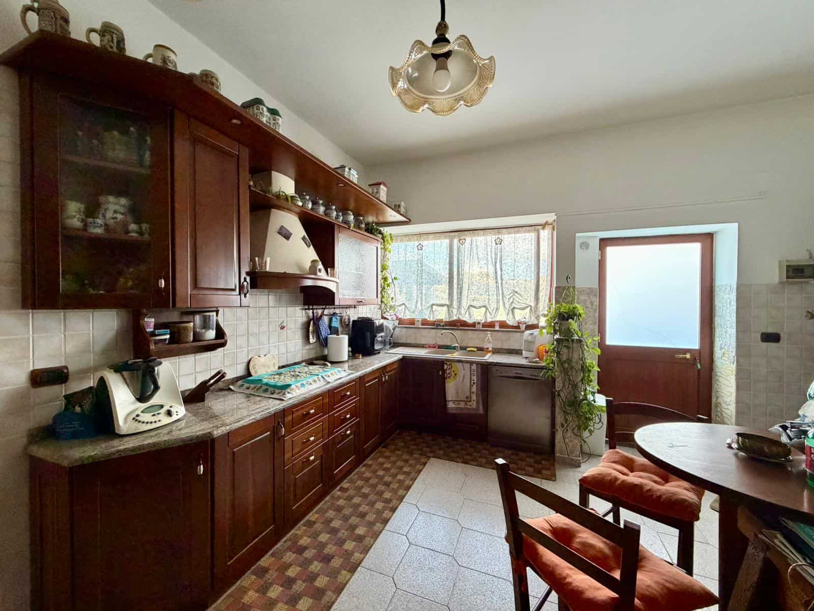 Lavagna da cucina con mobili in legno scuro, elettrodomestici e accessori, illuminata da luce naturale attraverso la finestra. Spazio funzionale e accogliente per preparare i pasti.