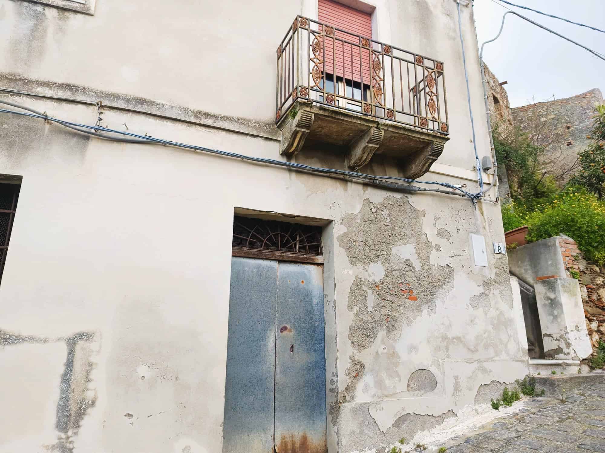 Porte in metallo arrugginito e balcone in ferro battuto su edificio storico con pareti in pietra e intonaco deteriorato, situato in zona collinare con strada acciottolata.