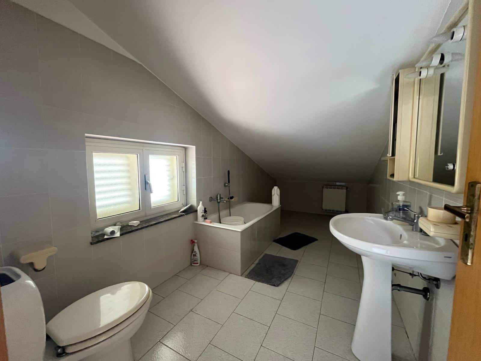 Lavabo e vasca da bagno in un bagno mansardato con pareti e pavimenti chiari, finestra con persiana, specchio e accessori bagno, ambiente luminoso e spazioso.