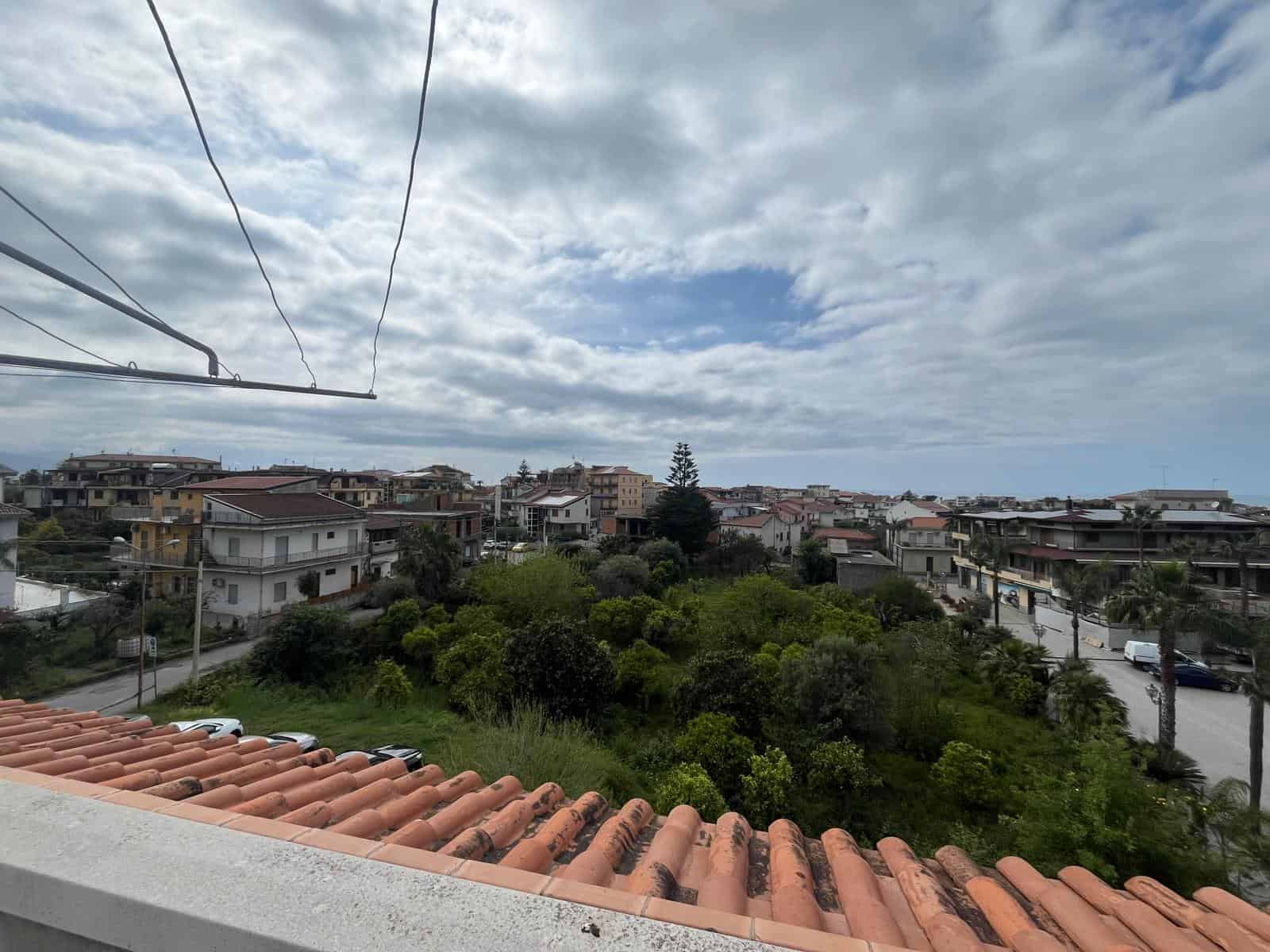 Vista panoramica di un quartiere residenziale con case e vegetazione, sotto un cielo nuvoloso, vista da un balcone o terrazza con tegole in terracotta.