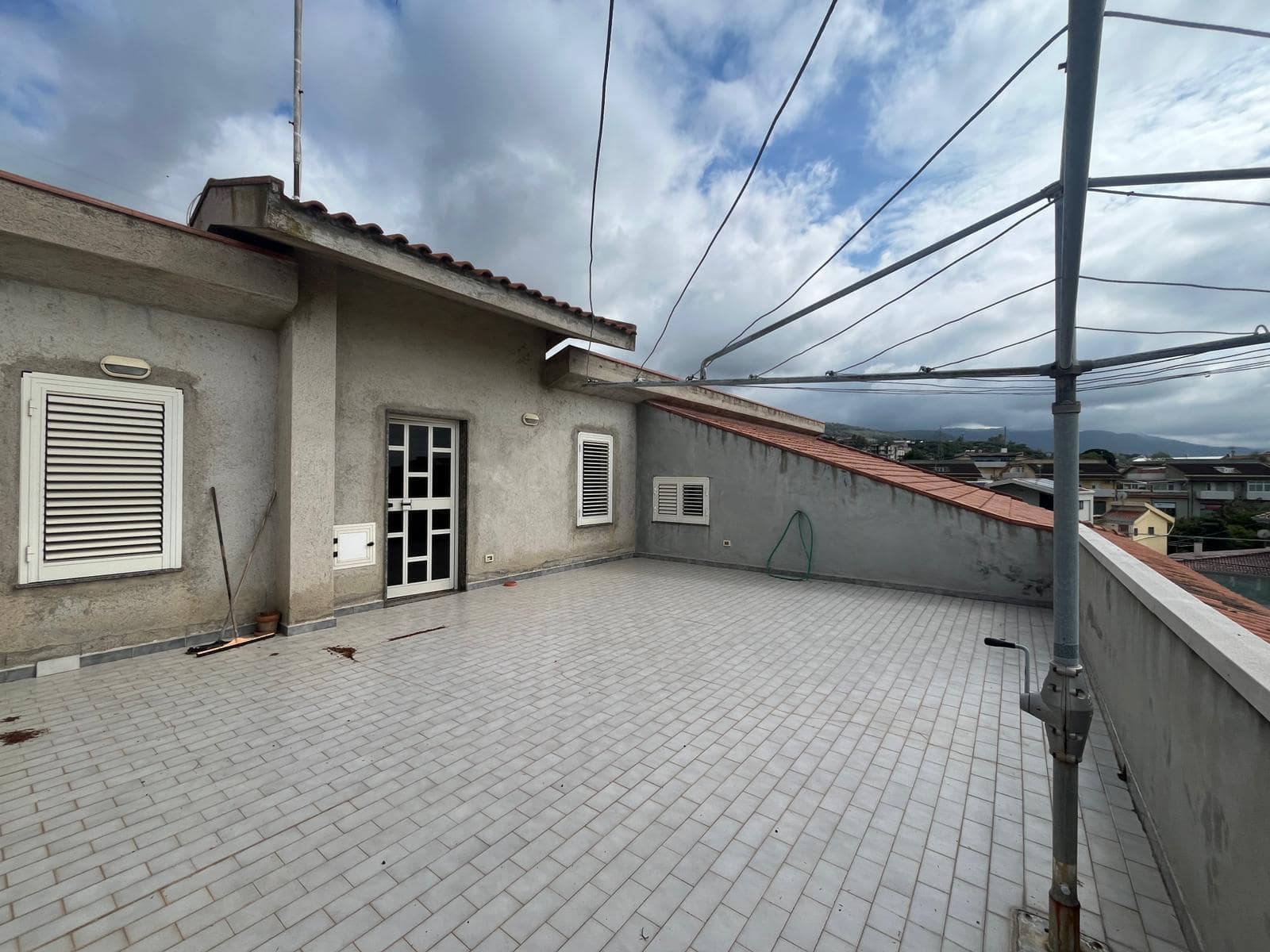 Balcone panoramico di un appartamento con vista sulla città, pavimentato in piastrelle grigie e parapetto in cemento, con linee di cavi elettrici e un cielo nuvoloso sullo sfondo.