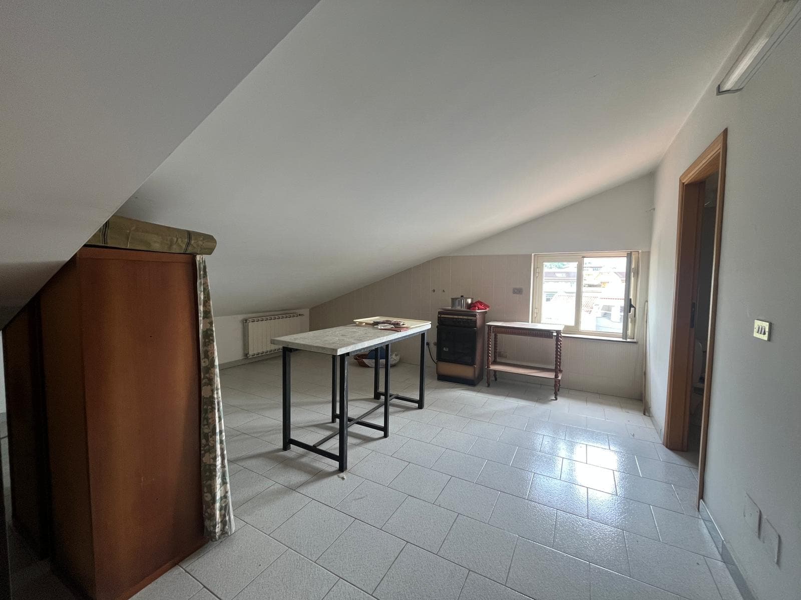 Ampia stanza mansardata con finestra, pavimento in piastrelle chiare, arredi minimalisti, ideale come studio o spazio living, luminoso e ben ventilato, perfetto per soluzioni di arredamento moderne e.