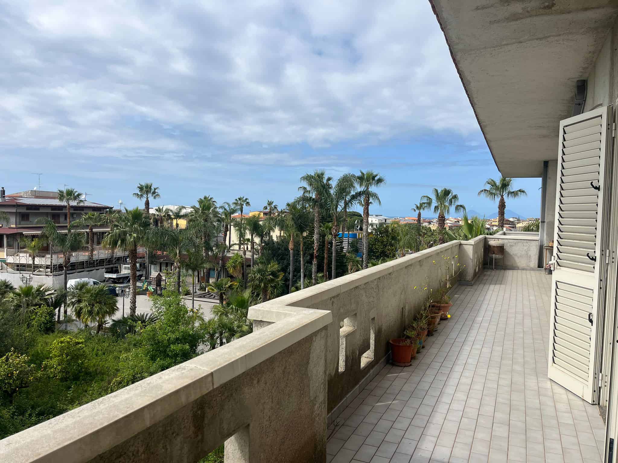 Balcone di un appartamento con vista su palme e il mare in lontananza, cielo nuvoloso, con piante in vaso lungo il parapetto.