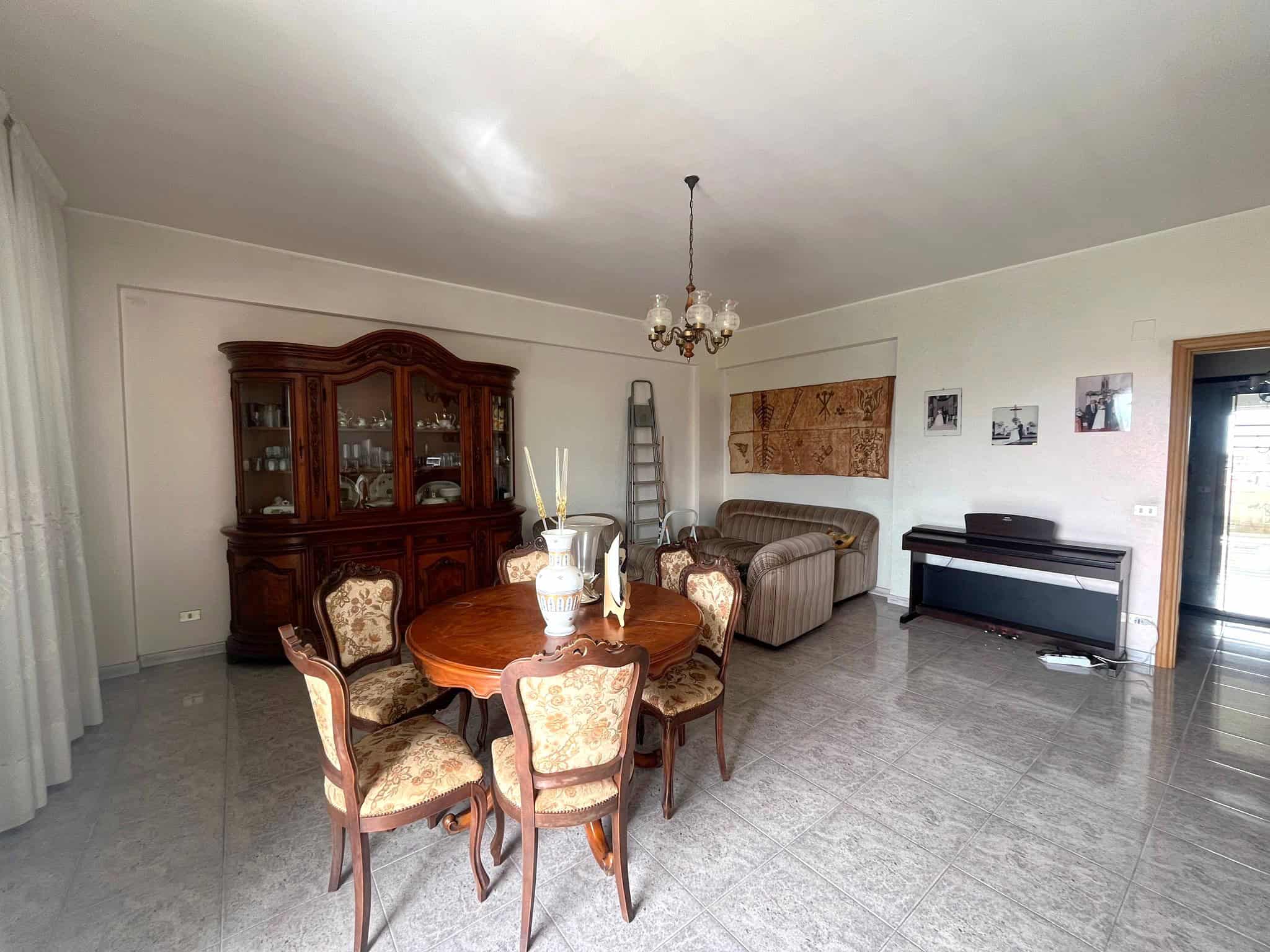 Elegante soggiorno con tavolo da pranzo in legno, sedie imbottite, grande credenza in stile classico, divano e pianoforte, ambiente luminoso e accogliente con decorazioni e fotografie alle pareti.