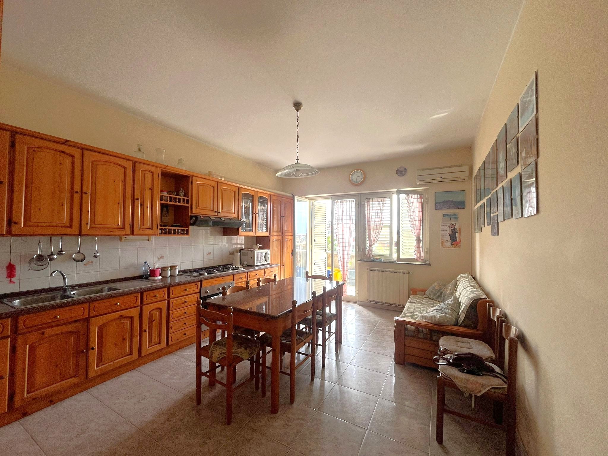 Ampia cucina abitabile con mobili in legno, tavolo centrale e zona soggiorno con divano, luminosa e accogliente, ideale per famiglie e incontri conviviali, in un appartamento ben arredato.