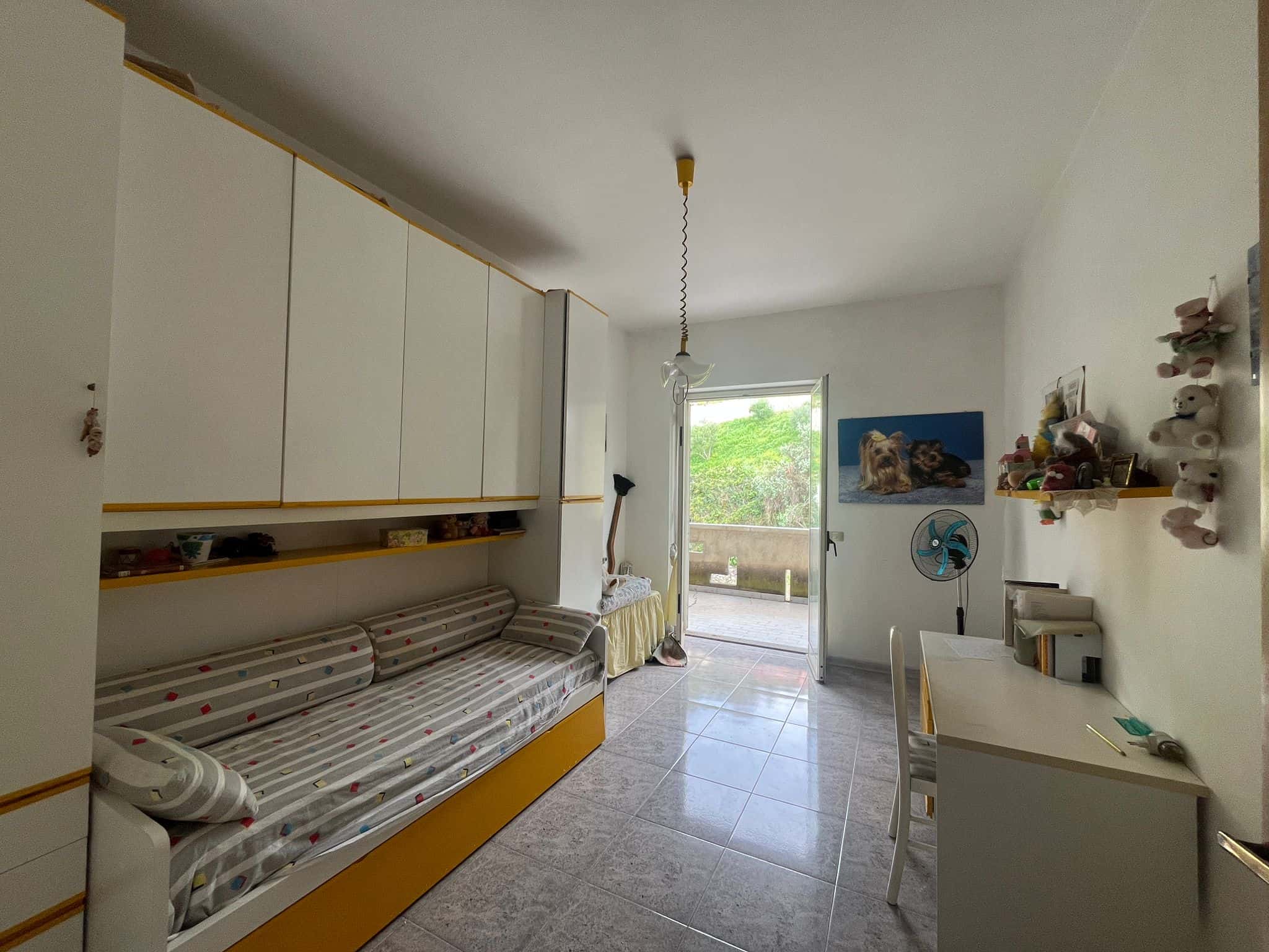 Il letto a castello in una camera da letto con accessori e decorazioni, vista verso il balcone con vista verde. Ideale per spazi familiari o camerette per bambini, combinando funzionalità e comfort.