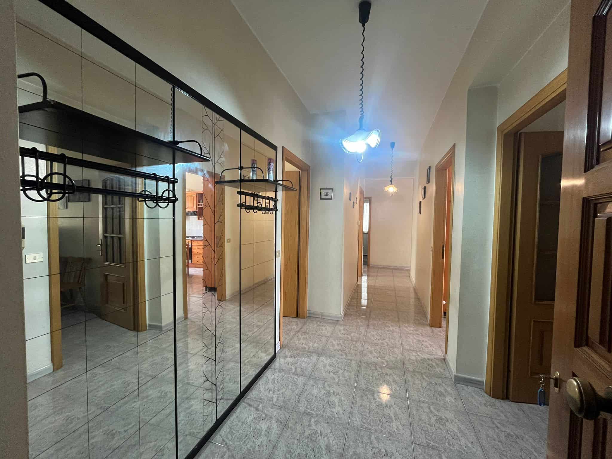 Ampio corridoio interno di un appartamento con pareti chiare, pavimento in piastrelle lucide e illuminazione soffusa, porte in legno naturale e specchi a parete che riflettono la luce.