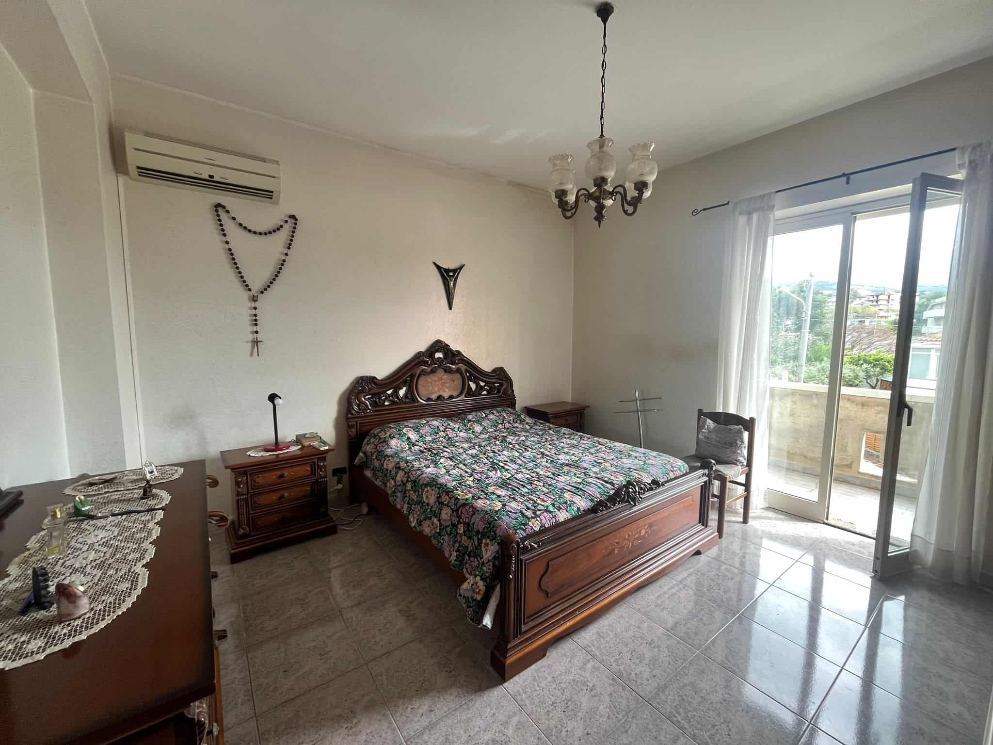 Letto matrimoniale in camera da letto con arredi classici e decorazioni religiose, luce naturale che entra dalla finestra, ambiente spazioso e accogliente.