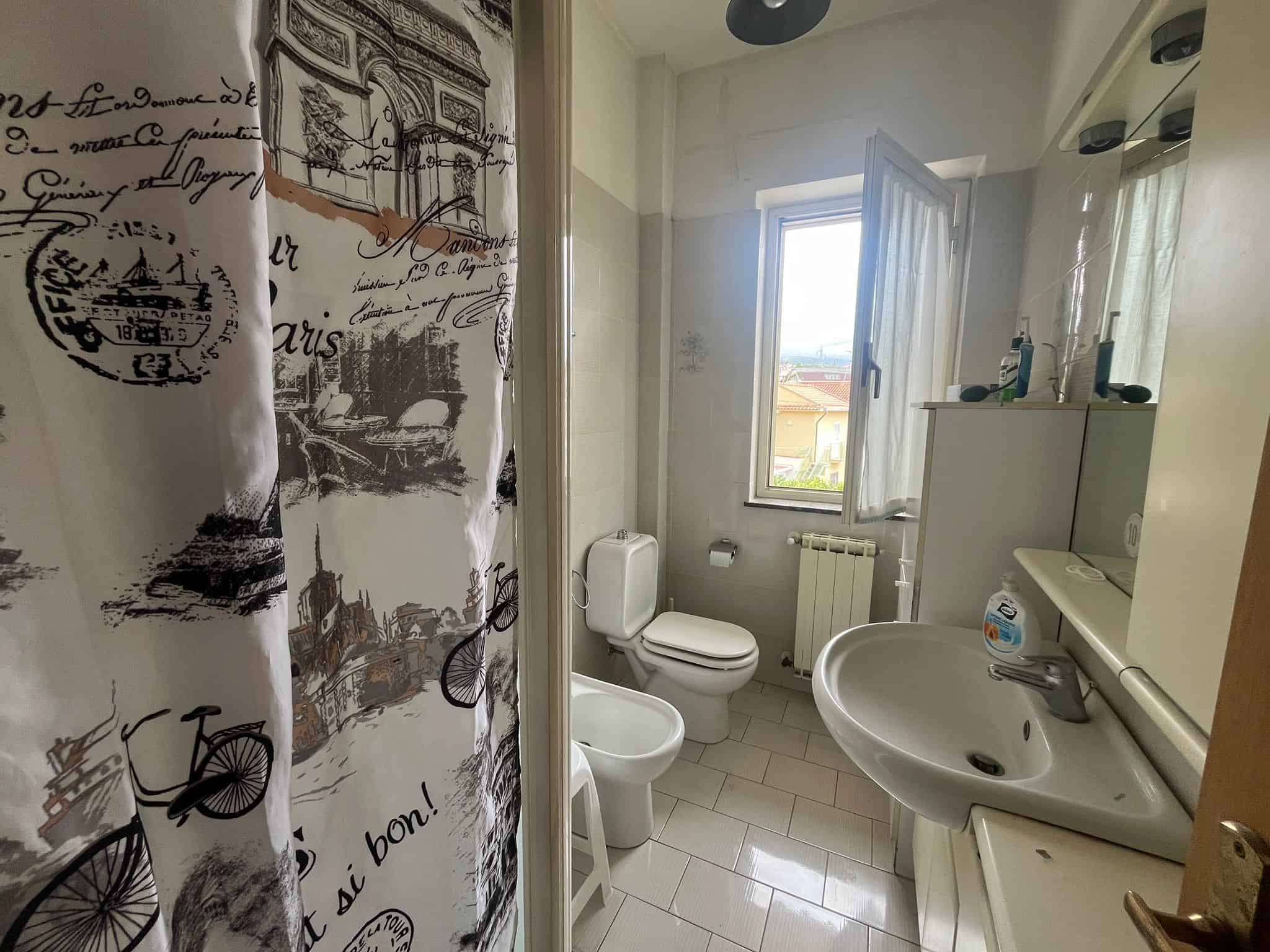 Doccia moderna con tende decorative in un bagno luminoso e arioso, dotato di finestra e sanitari essenziali, ideale per ristrutturazioni e miglioramenti domestici.