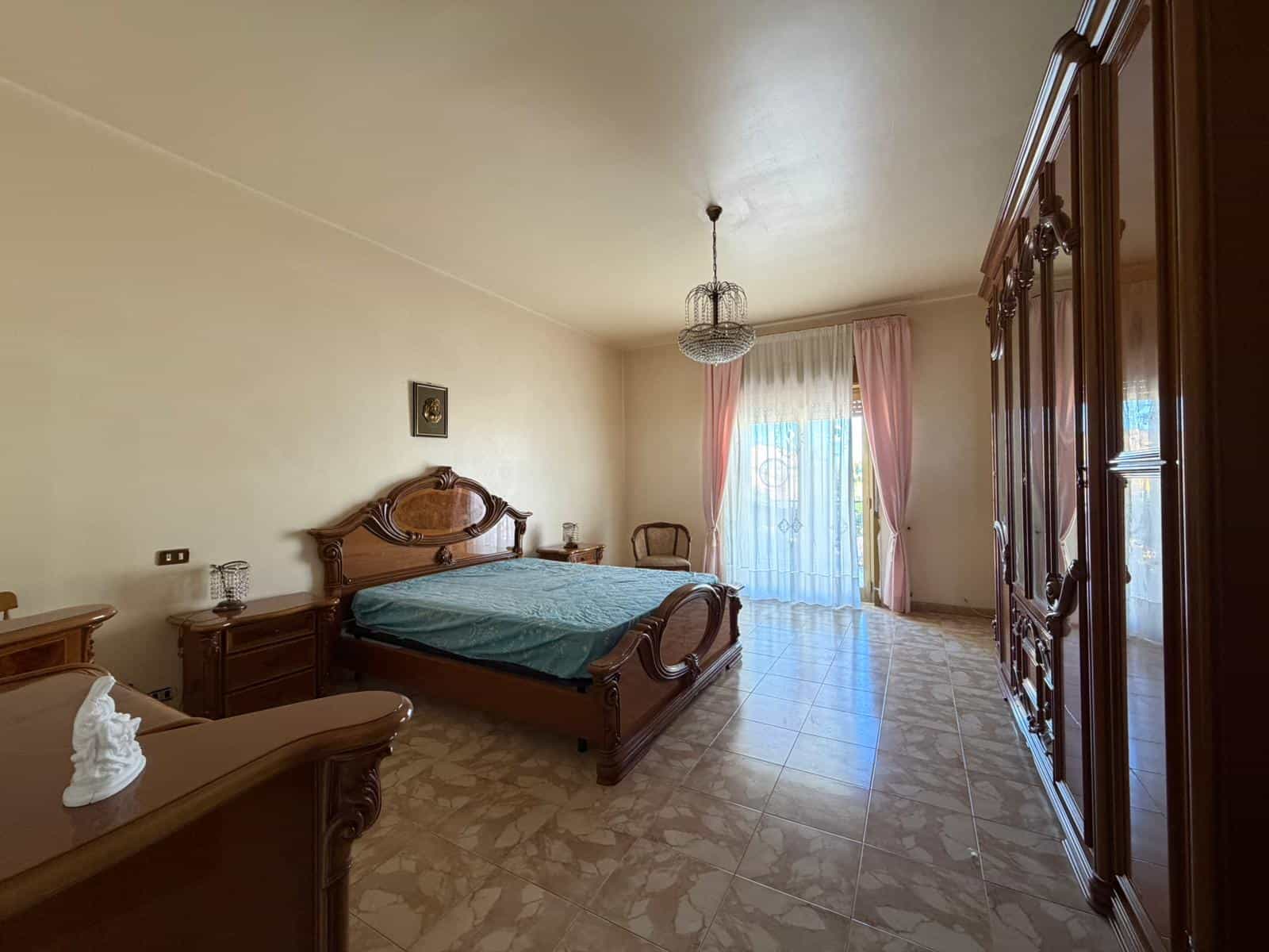 Ampia camera da letto con arredi in legno, grande armadio e finestra con tende rosa, ambiente luminoso e accogliente, ideale per affitti o vendita immobili di prestigio.