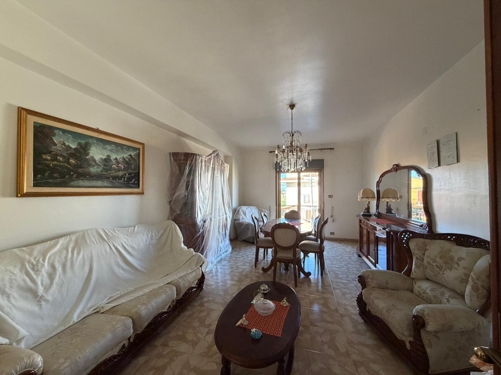 Ampia stanza soggiorno con mobili d'epoca, divani e tavolo da pranzo, illuminata da luce naturale e lampadario elegante, decorata con quadro paesaggistico e specchio antico, ambiente accogliente e raf.