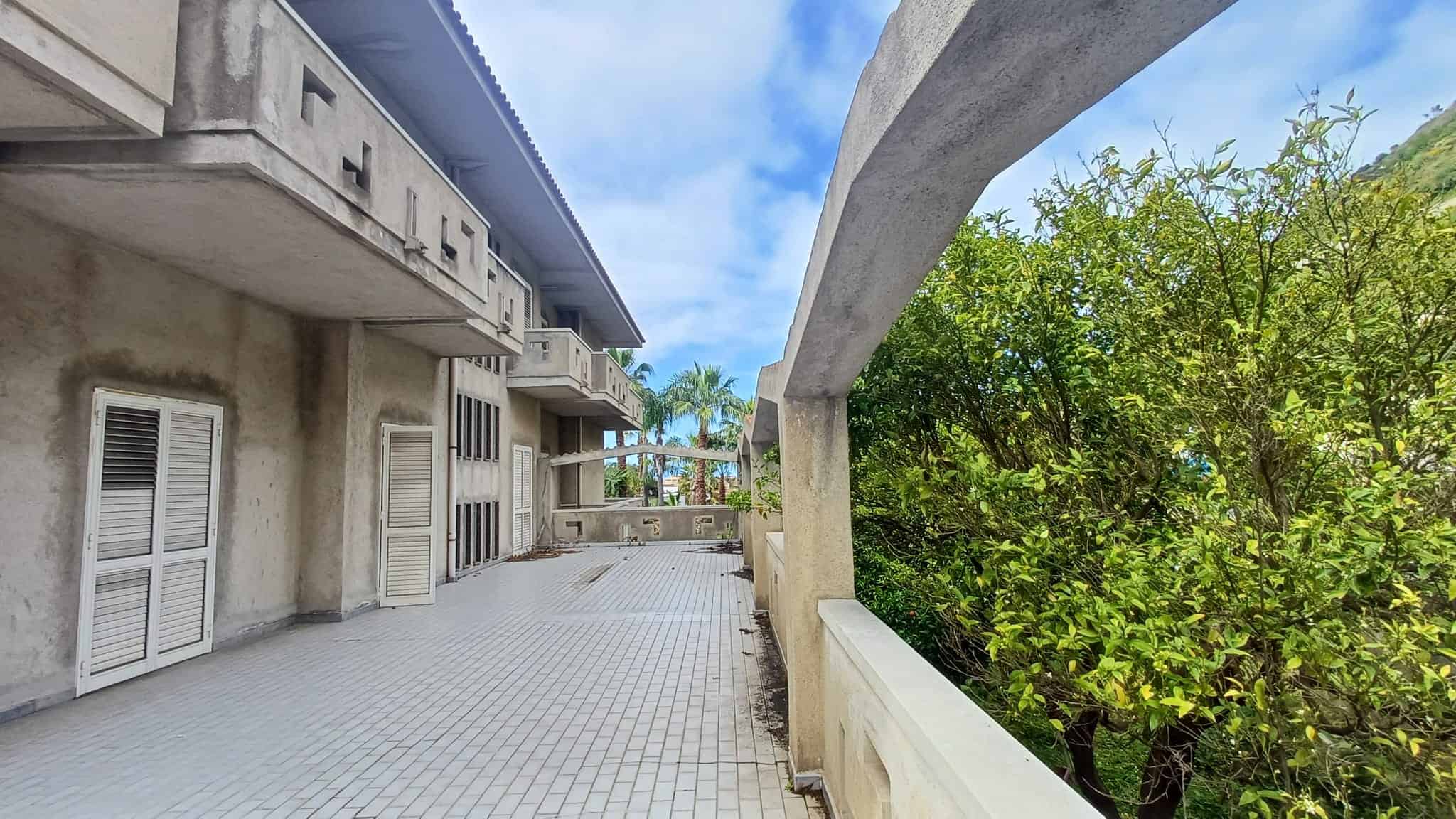 Moderno balcone con vista su giardino verde e edificio residenziale, in un complesso abitativo con struttura in cemento e pareti bianche, sotto un cielo azzurro con nuvole sparse.