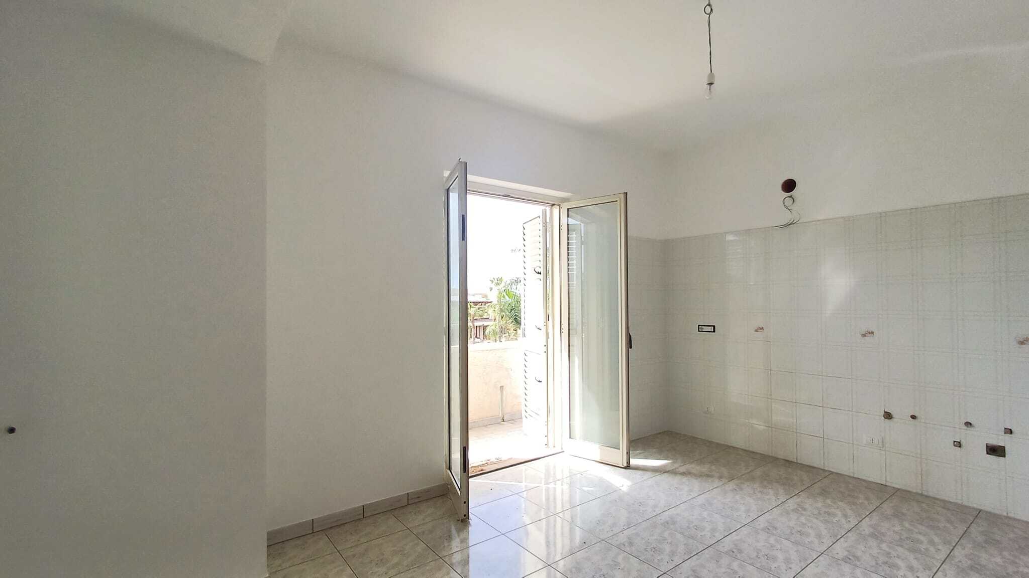 Porte finestra aperta che conduce a un balcone luminoso in una stanza vuota con pareti bianche e pavimento in piastrelle.