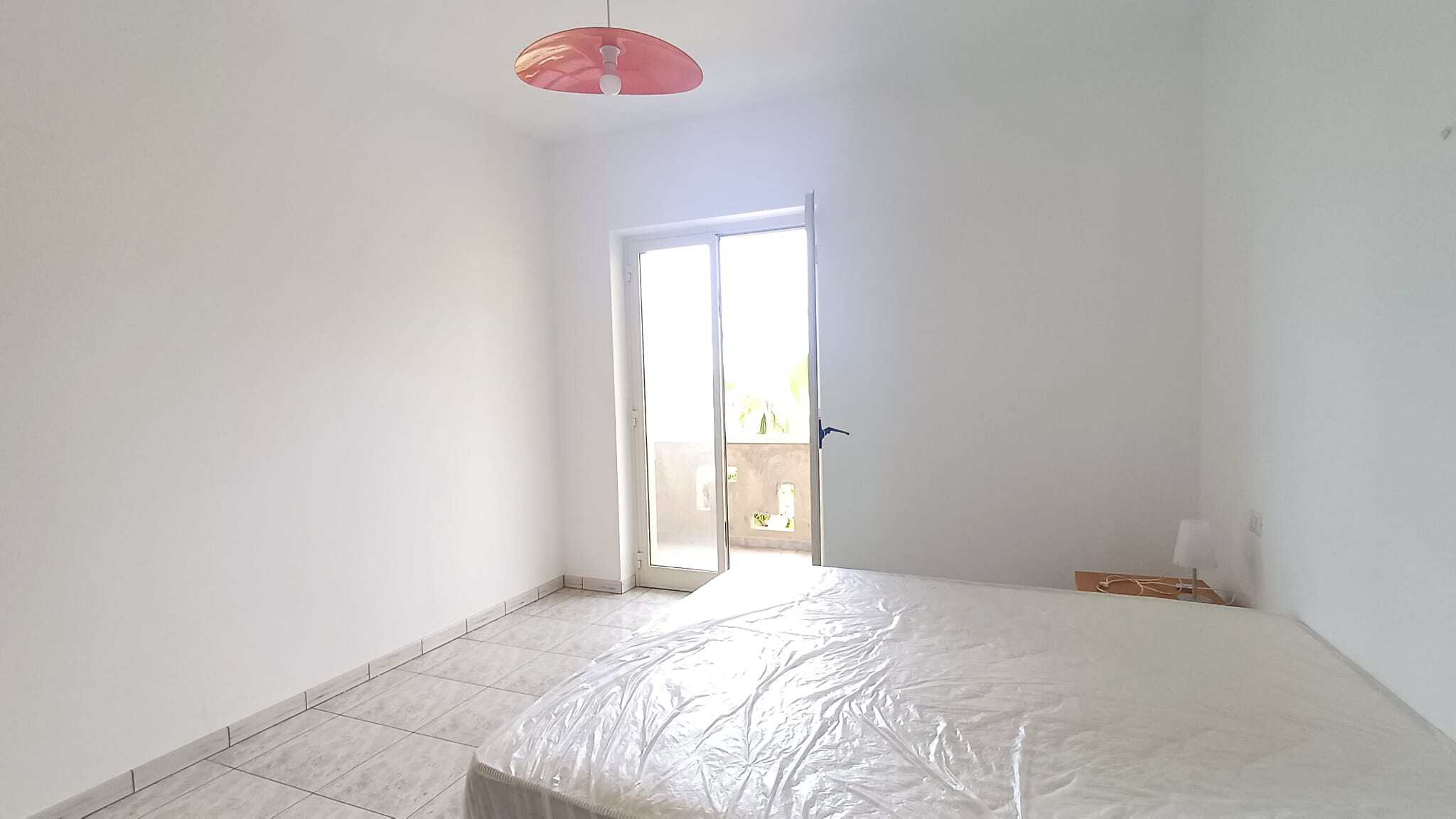 Ampia camera da letto luminosa con pareti bianche, grande finestra e porta finestra che permette l'accesso a un balcone, con soffitto con lampadario rosa e pavimento in piastrelle.