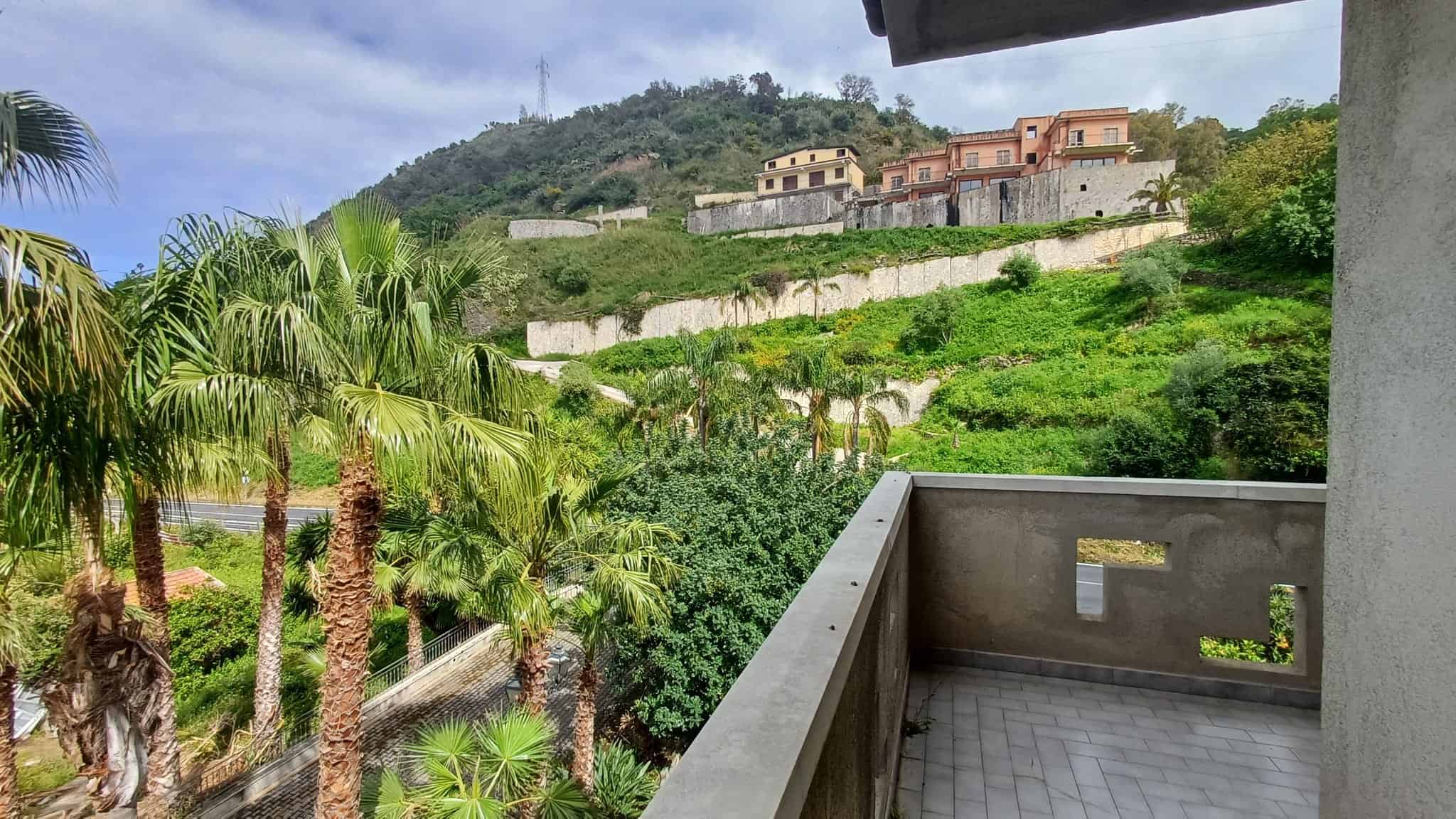 Balcone con vista panoramica su colline verdi e case in stile mediterraneo, circondato da palme e vegetazione lussureggiante, ideale per relax e momenti di tranquillità.