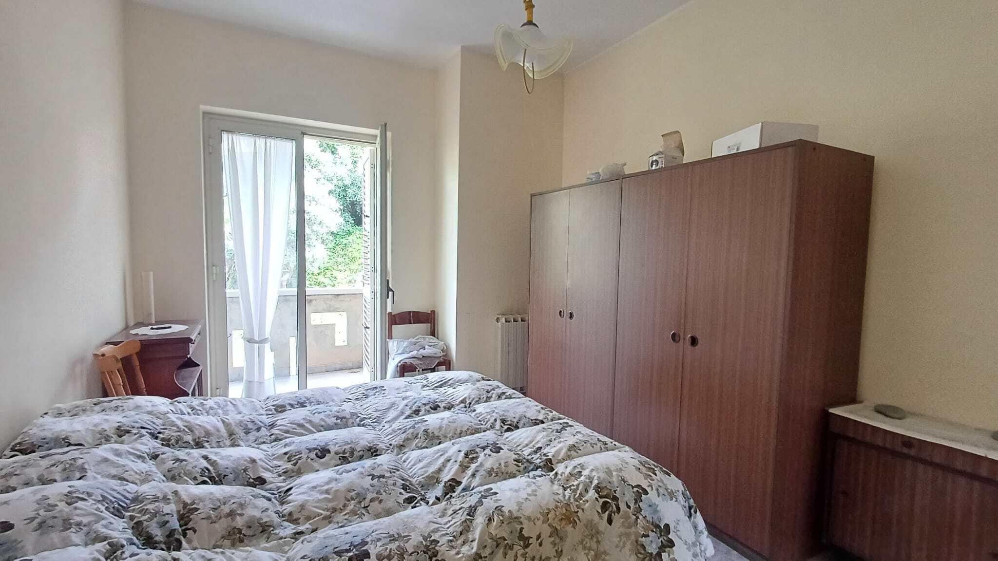 Ampia camera da letto con letto matrimoniale, armadio grande e accessori, luce naturale dalla porta finestra, arredamento semplice e funzionale, ambiente accogliente e luminoso.