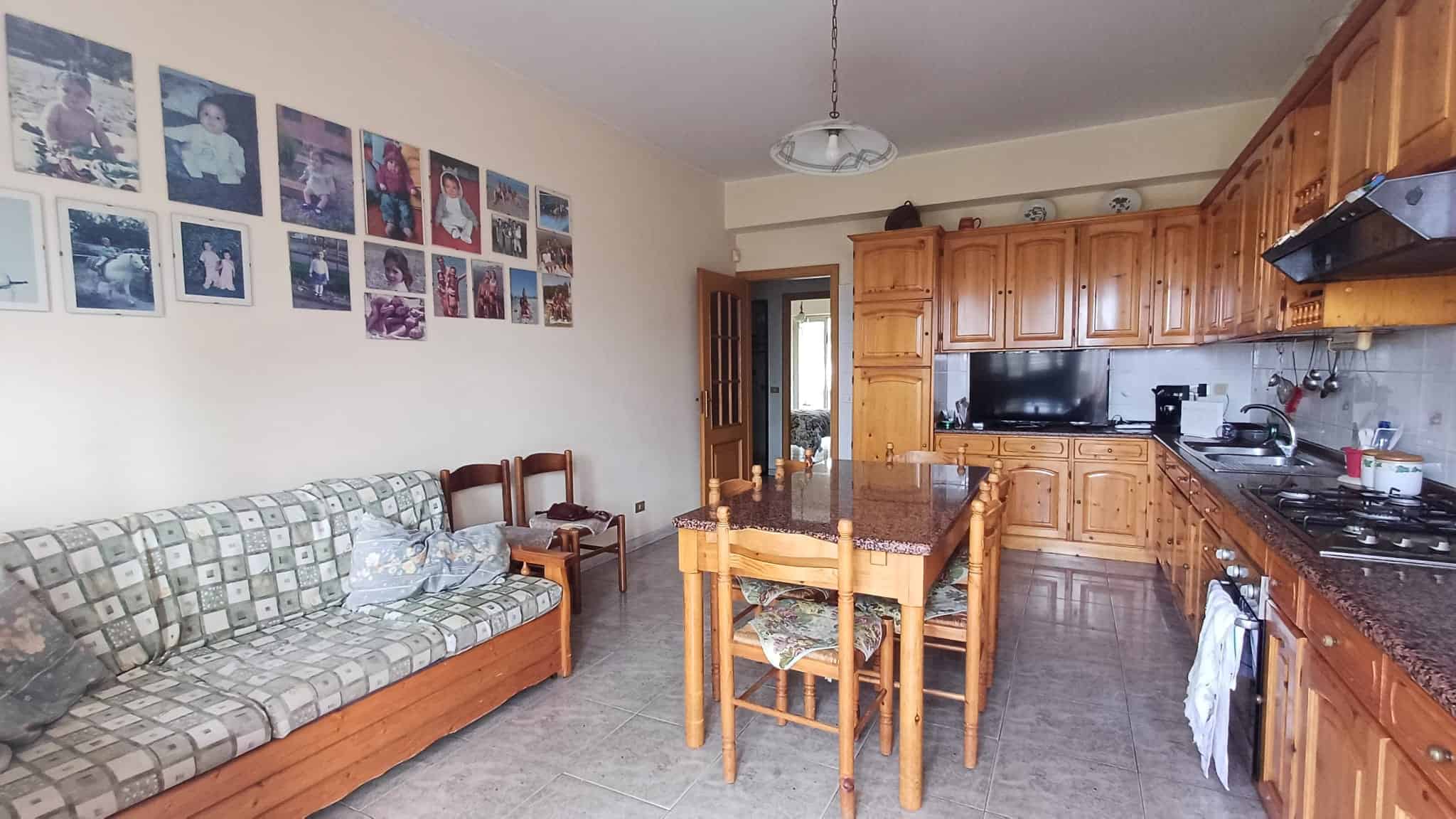 Cucina abitabile con mobili in legno, tavolo da pranzo e soggiorno con divano, decorata con foto alle pareti, ambiente luminoso e accogliente.