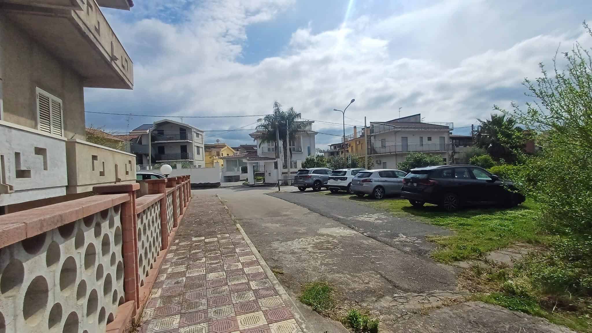 Ampio parcheggio esterno con auto parcheggiate, vicino a edifici residenziali e spazi verdi, sotto un cielo nuvoloso, in un'area urbana tranquilla e ben mantenuta.