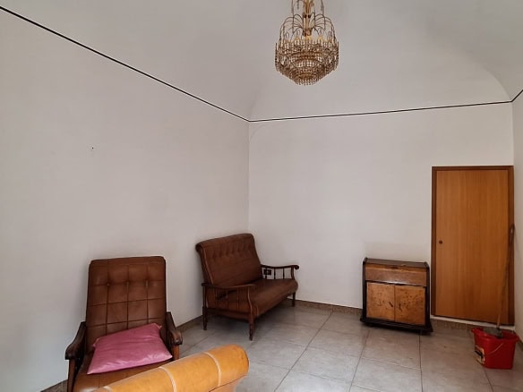 Ampia stanza con mobili vintage, pareti bianche e pavimento in piastrelle, arredi in legno e sedie imbottite, ideale per affitti o ristrutturazioni immobiliari.