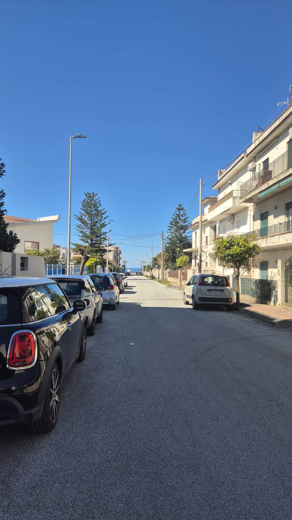Auto in strada residenziale con parcheggi, edifici moderni e alberi verdi sotto un cielo sereno, rappresentando un quartiere tranquillo e ben curato.