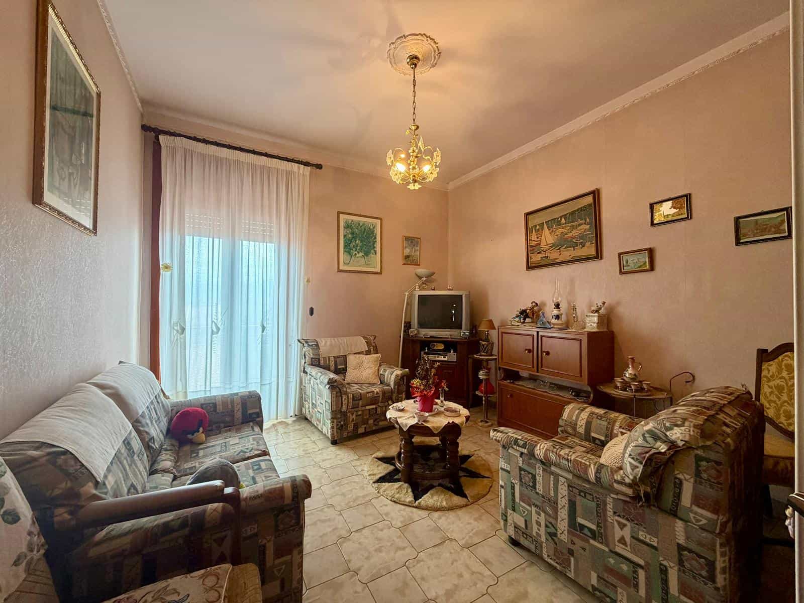 Una stanza soggiorno classica con divani e poltrone, televisore e decorazioni alle pareti, illuminata da una lampada a soffitto e luce naturale dalla finestra con tende bianche.