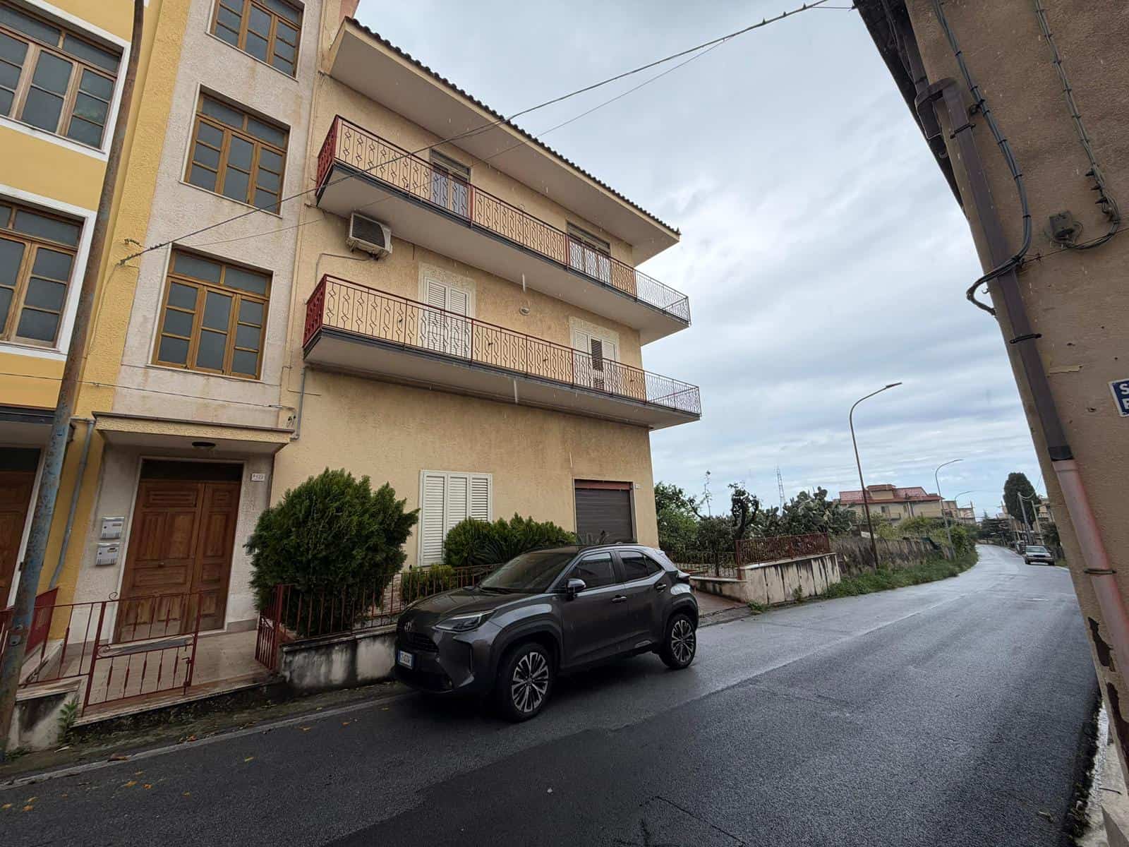 Edificio residenziale con tre piani e balconi, situato in una zona urbana, con auto parcheggiata davanti e strada asfaltata.