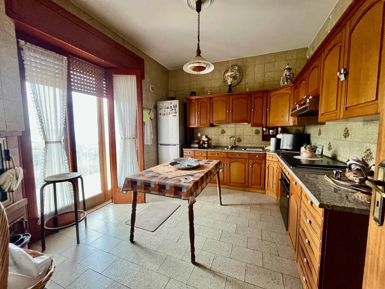 Cucina abitabile con mobili in legno, tavolo centrale e accessori, luce naturale da porta finestra, ambiente accogliente e funzionale.