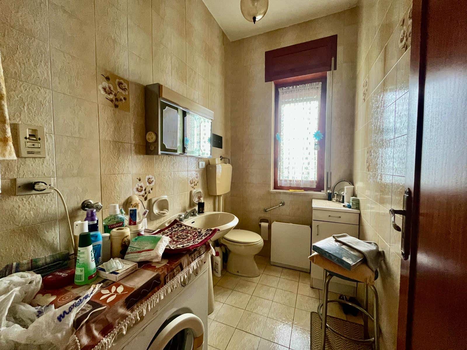 Doccia e servizi igienici in un bagno di piccole dimensioni con pareti rivestite di piastrelle beige, finestra con tende, e vari oggetti di uso quotidiano disposti sul mobile e sul piano del lavabo.